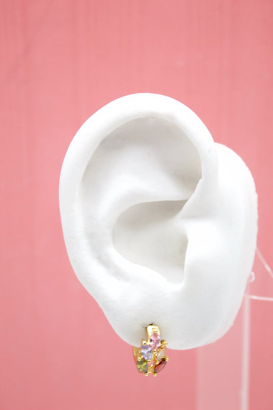 A white mannequin ear displays a gold huggie hoop earring adorned with multicolored gemstone charms against a pink background.