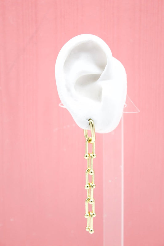 A white mannequin ear displays a modern gold paperclip chain dangle earring against a solid pink backdrop.