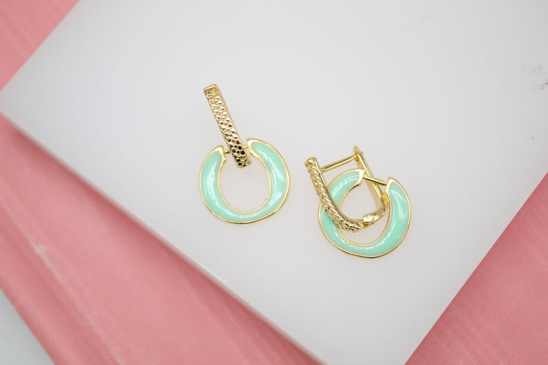 A pair of gold huggie hoop earrings featuring mint green enamel and textured bars rest on a white surface.