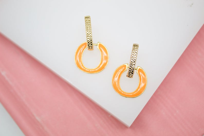 A pair of gold earrings featuring textured rectangular posts and dangling orange enamel hoops rests on a white and pink surface.