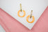 A pair of gold earrings featuring textured rectangular posts and dangling orange enamel hoops rests on a white and pink surface.