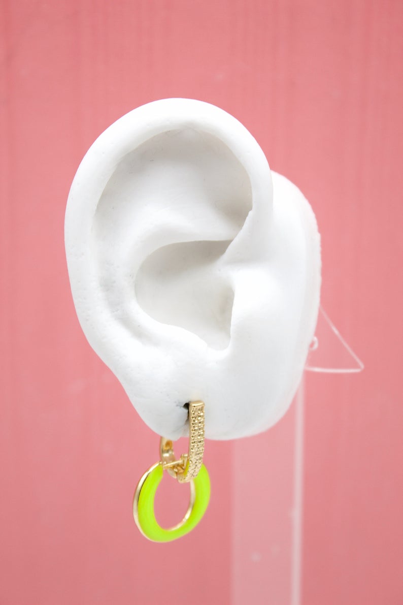 A white mannequin ear displays a gold hoop earring accented with a vibrant neon green ring against a solid pink backdrop.