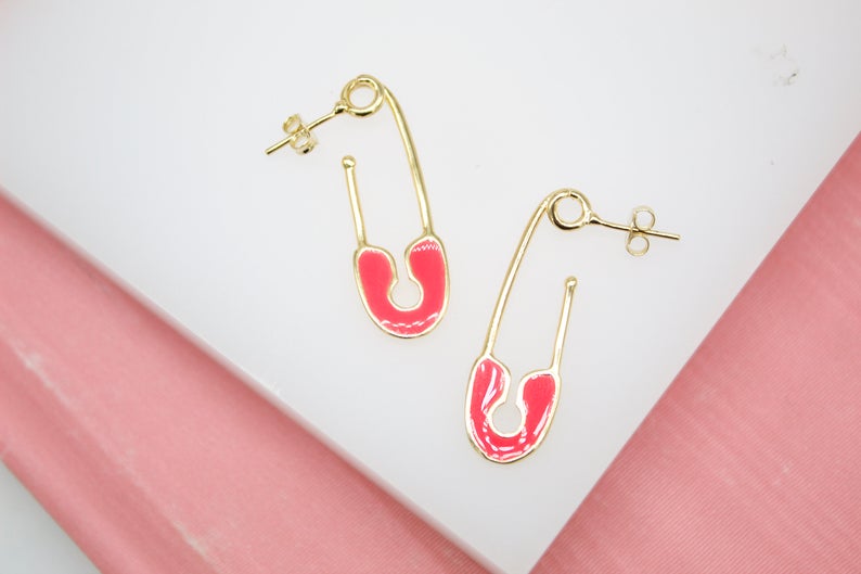 A pair of gold safety pin earrings featuring vibrant hot pink enamel details rest on a white surface against a pink background.