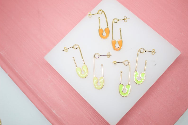 Six gold safety pin earrings with colorful enamel accents rest on a white display surface against a pink background.
