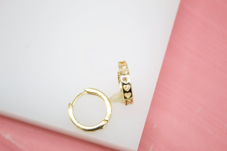 A pair of gold huggie hoop earrings featuring cutout heart details rests on a white and pink display surface.
