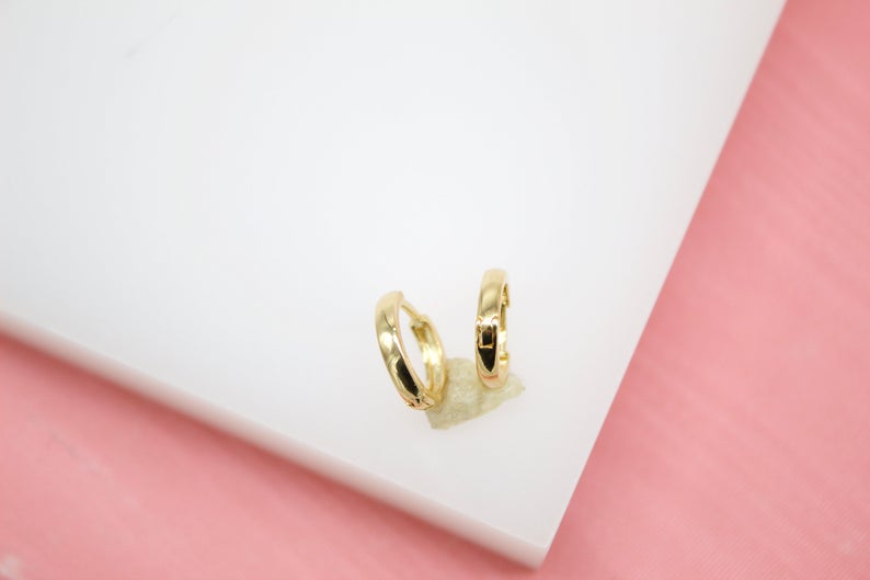 A pair of polished gold huggie hoop earrings rests delicately on a white display stand.