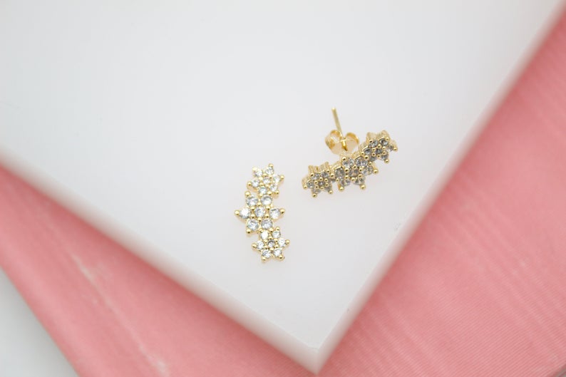 A pair of gold floral cluster stud earrings with sparkling clear cubic zirconia stones rests elegantly on a white and pink surface.
