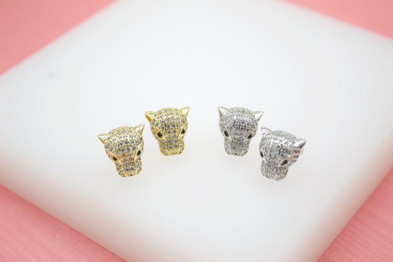 Two pairs of sparkling panther head stud earrings, one in gold and one in silver, rest on a white display surface against a pink background.
