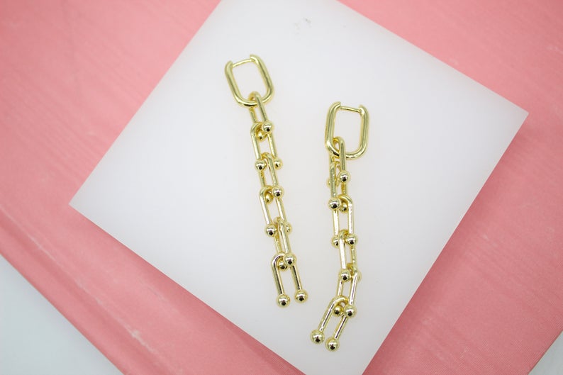 A pair of gold chain-link dangle earrings featuring geometric links and small bead details rests on a white display surface against a pink background.