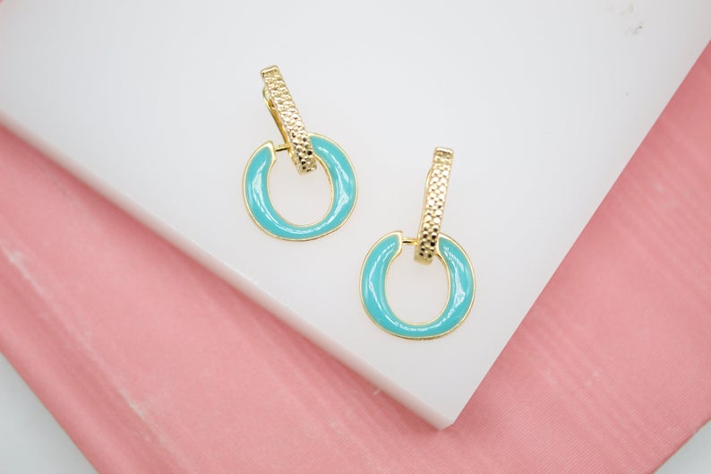 A pair of gold and turquoise enamel drop hoop earrings rests on a white and pink display surface.