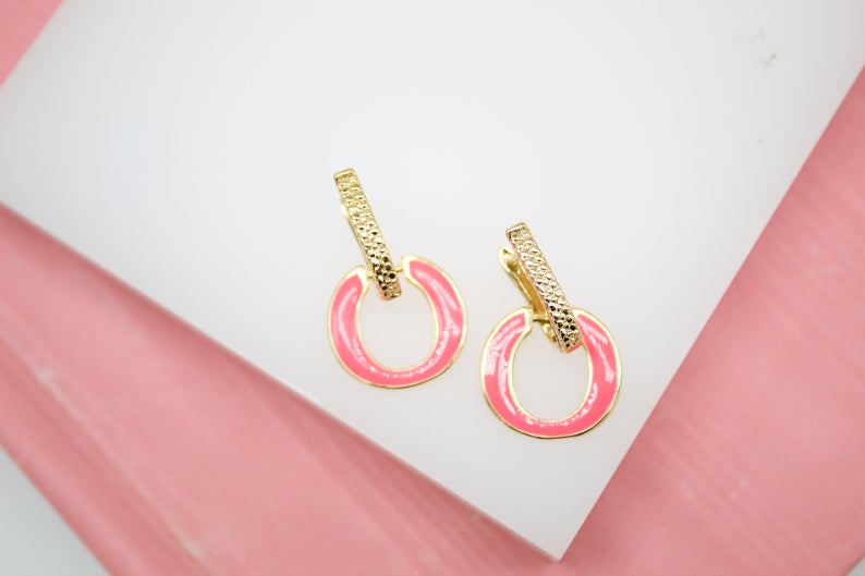 A pair of gold earrings featuring textured rectangular posts and bright pink enameled hoops rest on a white display surface.