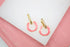 A pair of gold earrings featuring textured rectangular posts and bright pink enameled hoops rest on a white display surface.