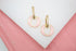 A pair of gold huggie earrings featuring pink enamel circular drops rests on a white display surface.