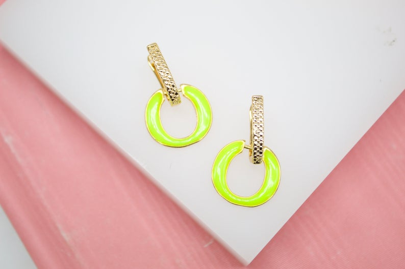 A pair of gold earrings featuring textured rectangular posts and bright neon green enamel hoops rests on a white display surface.