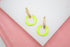 A pair of gold earrings featuring textured rectangular posts and bright neon green enamel hoops rests on a white display surface.