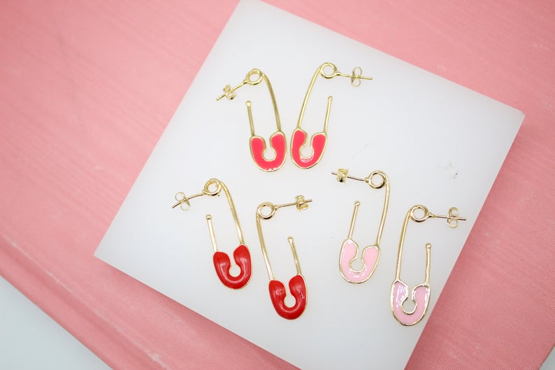 Six gold safety pin earrings with enamel details in red and light pink display neatly on a white surface against a pink background.