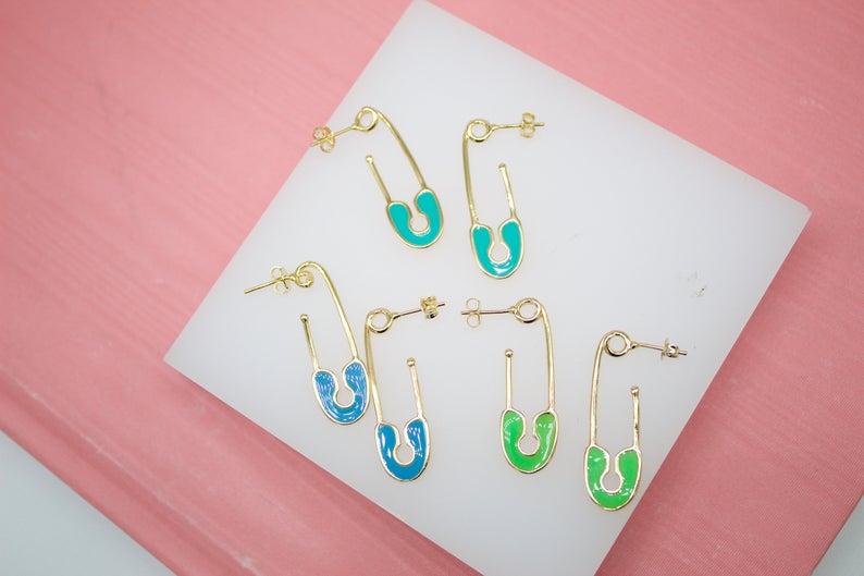 Six gold safety pin earrings in turquoise, blue, and green enamel display on a white and pink surface.