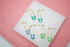 Six gold safety pin earrings in turquoise, blue, and green enamel display on a white and pink surface.