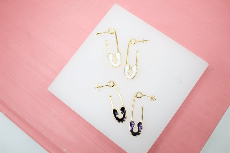 Four gold safety pin earrings, two with white enamel and two with dark purple enamel, rest on a white display tile against a pink background.