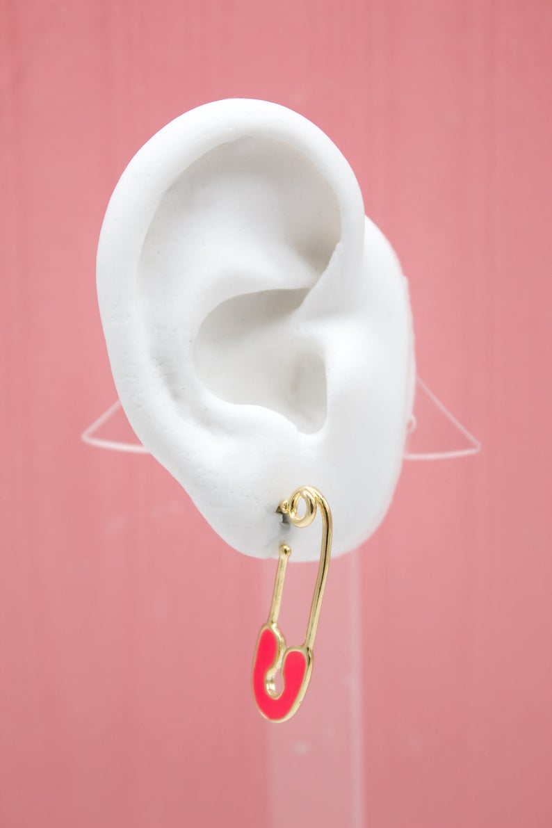 A white mannequin ear displays a gold safety pin earring featuring a vibrant pink enamel accent against a soft pink background.