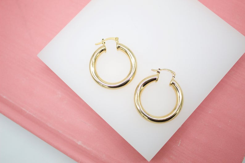 A pair of polished gold hoop earrings rests neatly on a white display surface against a pink background.
