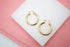 A pair of polished gold hoop earrings rests neatly on a white display surface against a pink background.