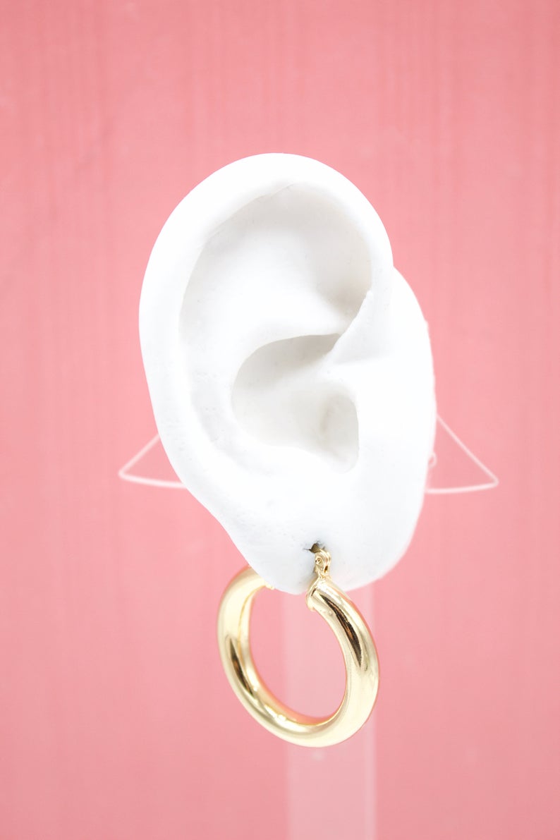 A white mannequin ear displays a thick gold hoop earring against a pink background.