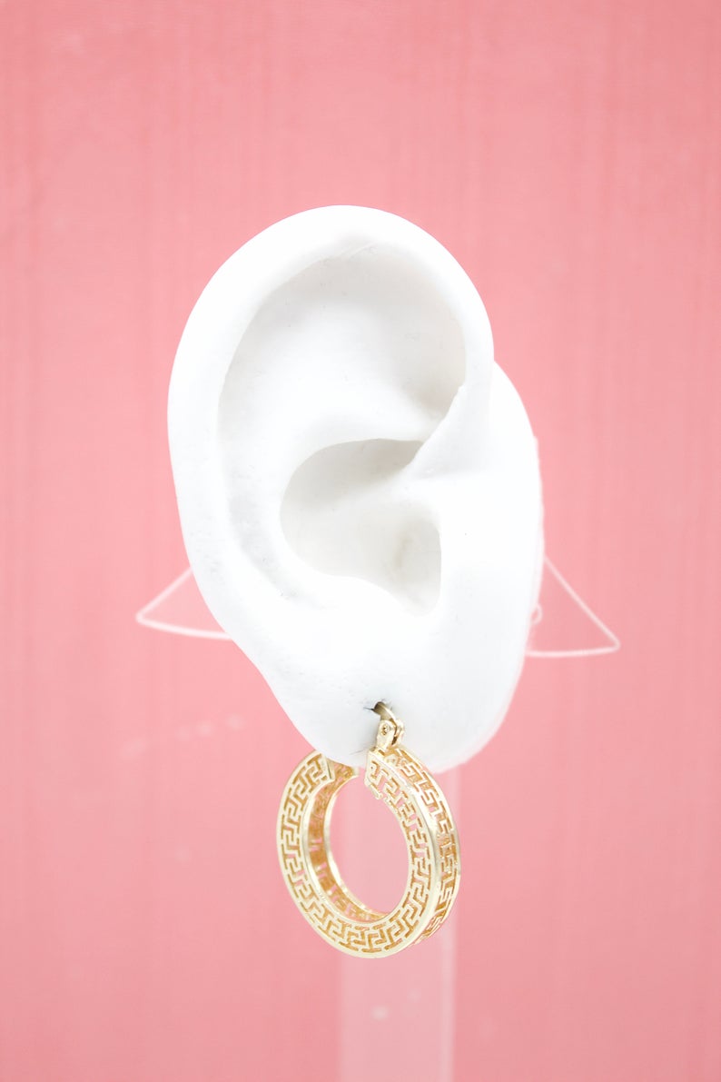 A white mannequin ear displays a gold Greek key hoop earring against a pink background.
