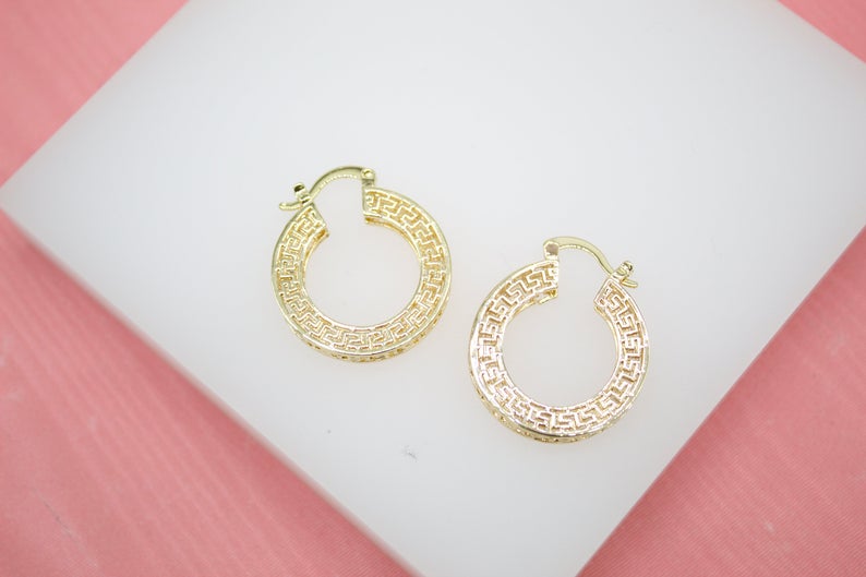 A pair of gold Greek key hoop earrings rests on a white display stand.