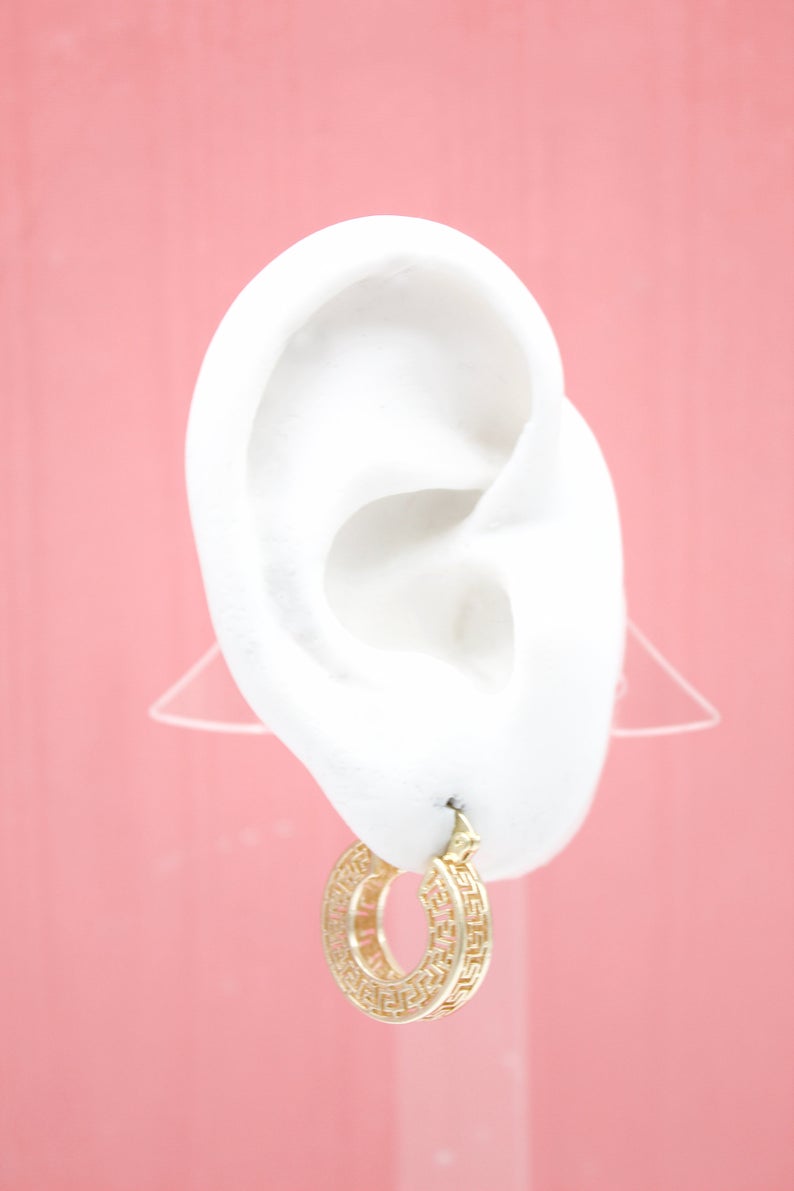 A white mannequin ear displays a thick gold hoop earring featuring an intricate Greek key pattern against a soft pink background.