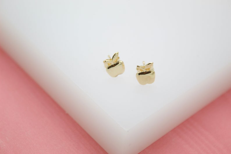 A pair of gold apple stud earrings rests on a white display block against a soft pink background.