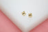 A pair of gold apple stud earrings rests on a white display block against a soft pink background.