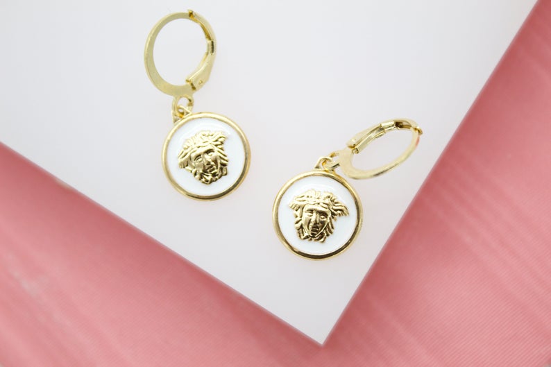 A pair of gold-plated hoop earrings featuring white enamel pendants with a raised Medusa head design rest on a white and pink display surface.