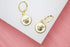 A pair of gold-plated hoop earrings featuring white enamel pendants with a raised Medusa head design rest on a white and pink display surface.