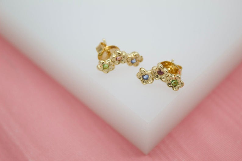 A pair of gold floral cluster stud earrings featuring sparkling multi-colored cubic zirconia stones rests on a white display surface.