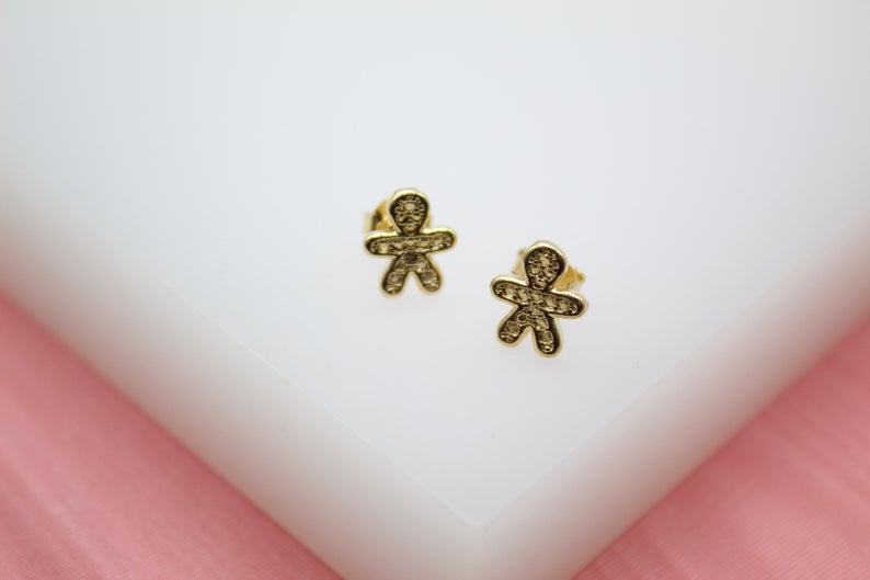 A pair of gold gingerbread man stud earrings featuring sparkling pave crystals rests on a white display stand.