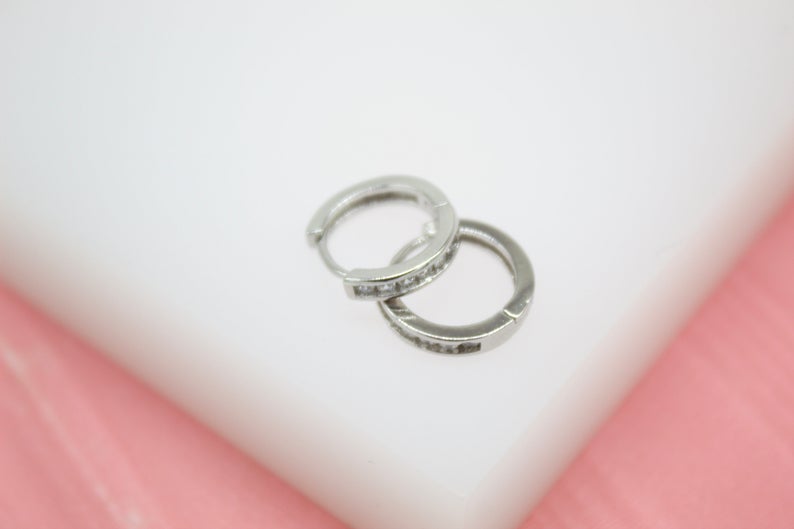 A pair of silver huggie hoop earrings featuring sparkling pave crystals rests on a white display surface.