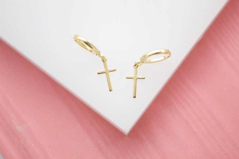 A pair of dainty gold cross hoop earrings rests on a white display surface against a pink background.