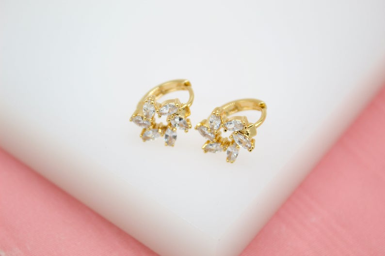 A pair of gold huggie hoop earrings featuring sparkling cubic zirconia stones arranged in a floral pattern rests on a white display surface.
