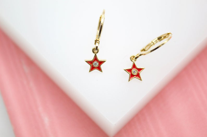 A pair of gold star hoop earrings featuring vibrant red enamel and a sparkling center stone rests on a white display against a pink background.