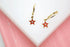 A pair of gold star hoop earrings featuring vibrant red enamel and a sparkling center stone rests on a white display against a pink background.