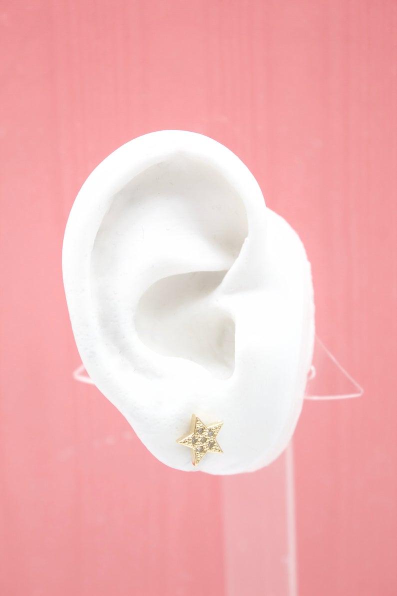 A white mannequin ear displays a sparkling gold star stud earring against a pink background.