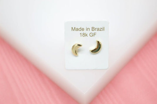 A pair of 18k gold-filled crescent moon stud earrings is displayed on a white display card with text that reads "Made in Brazil."