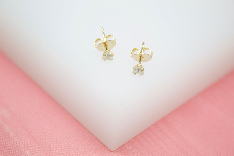 A pair of delicate gold stud earrings with sparkling cubic zirconia stones rests on a white and pink display surface, showcasing their secure backs.