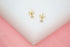 A pair of delicate gold stud earrings with sparkling cubic zirconia stones rests on a white and pink display surface, showcasing their secure backs.
