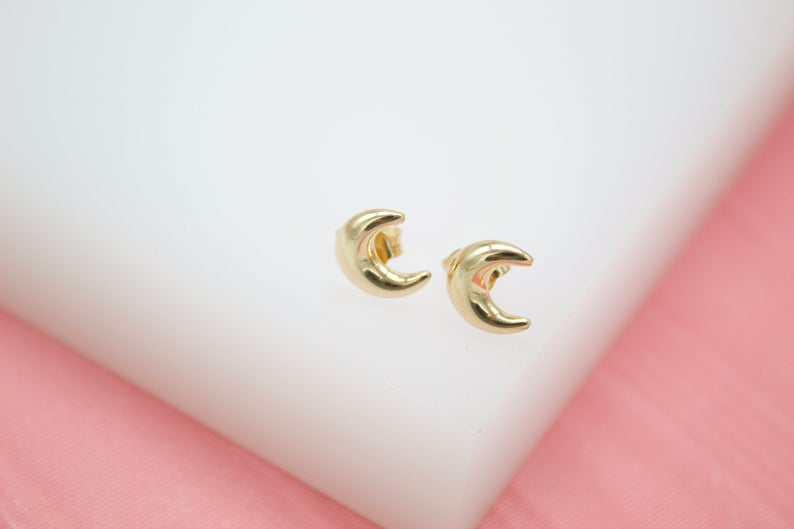 A pair of shiny gold crescent moon stud earrings rest on a white display surface against a pink background.