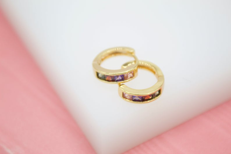 A pair of gold huggie hoop earrings featuring multicolored cubic zirconia stones rests on a white and pink surface.
