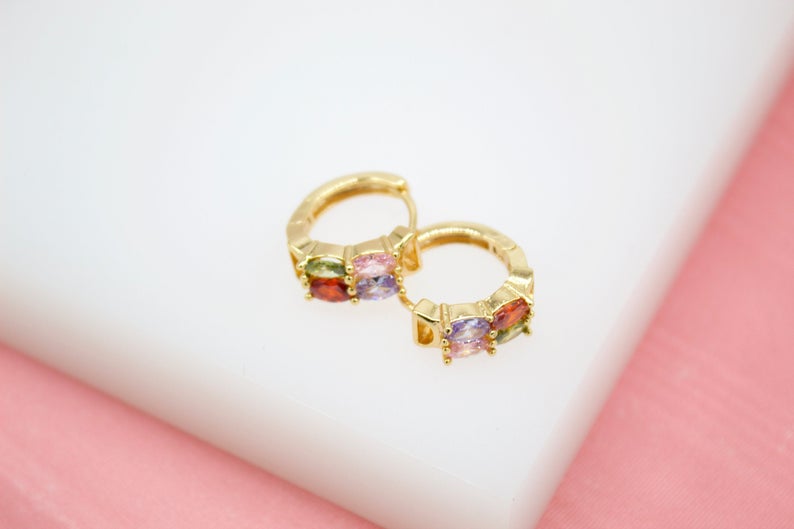 A pair of gold huggie hoop earrings features colorful multi-gemstone baguettes resting on a white display surface.