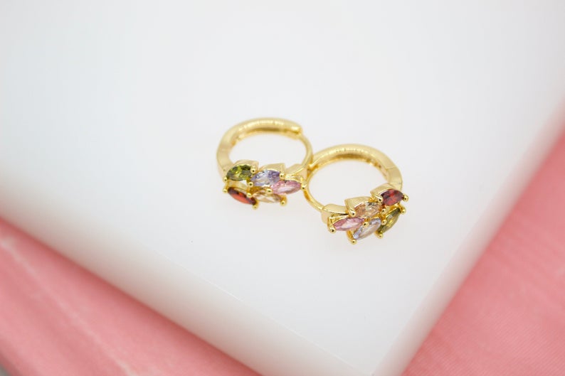 A pair of gold huggie hoop earrings featuring clusters of multicolored marquise-cut cubic zirconia stones rests on a white display surface.