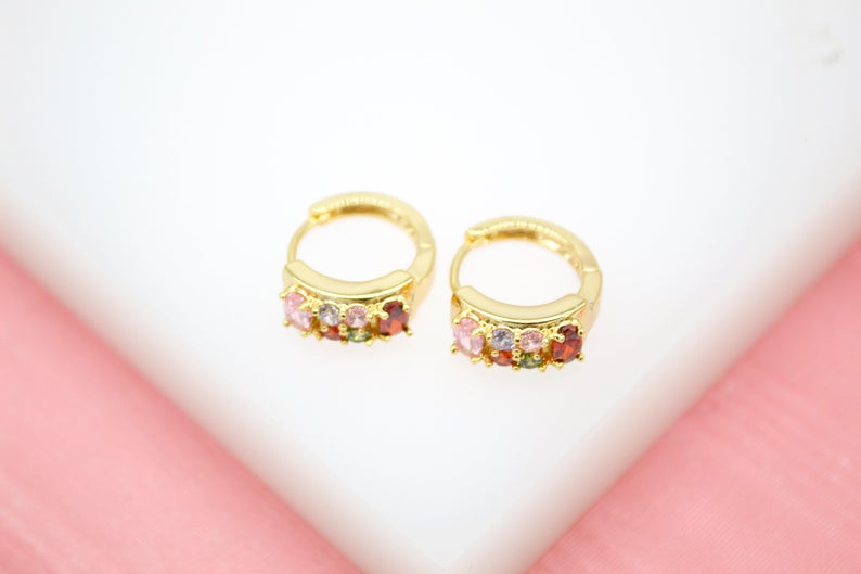 A pair of gold huggie hoop earrings embedded with colorful multi-gemstones rests on a white display surface against a pink background.